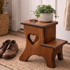 以下が含まれることがあります： ハート型の切り抜きがある2段の木製スツール。スツールは温かみのある茶色に着色されており、上に緑の植物が入った小さな白いセラミックポットがあります。茶色の革靴がジュートラグの上にあります。