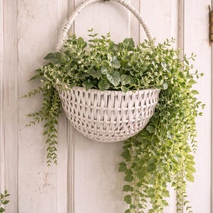 Könnte beinhalten: Ein weißer Weidenkorb, gefüllt mit grün-weißen Rankpflanzen, hängt an einem Messinghaken an einer weißen Tür. Der Korb hat ein gewebtes Design und einen gebogenen Griff. Die Pflanzen fallen an den Seiten herab.