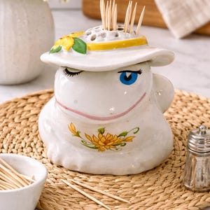May include: A whimsical white ceramic toothpick holder shaped like a smiling face wearing a hat. The hat is decorated with a yellow band and a sunflower. The holder is filled with wooden toothpicks. A small bowl of toothpicks is also visible.