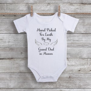 White baby bodysuit on a gray, plank background. Text in black with silver angel wings and halo. Text reads Hand Picked for earth by my great dad in heaven.