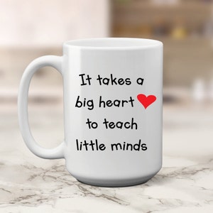 May include: White ceramic coffee mug with a black inscription that reads "It takes a big heart to teach little minds" with a red heart symbol.
