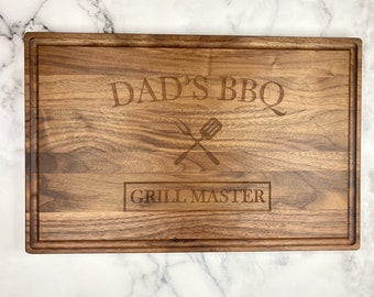 May include: A brown wooden cutting board with the text "Dad's BBQ Grill Master" engraved on it.