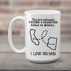 Large white mug, with handle. The text The love between father & daughter knows no distance is at the top. An outline of 2 states is below connected with a dotted line. Blue hearts are placed in each state.  The text I love you dad is below.