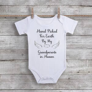 May include: White baby onesie with black text that reads "Hand Picked For Earth By My Grandparents in Heaven". The text is surrounded by a pair of silver glitter angel wings.