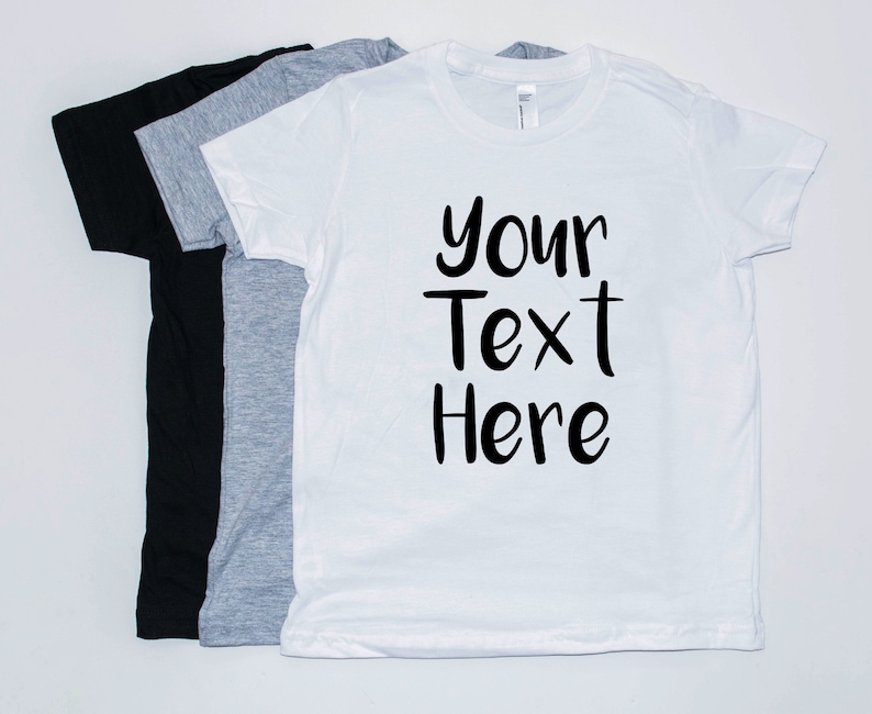 May include: Three t-shirts, one black, one gray, and one white. The white t-shirt has the text "Your Text Here" printed on it in black.