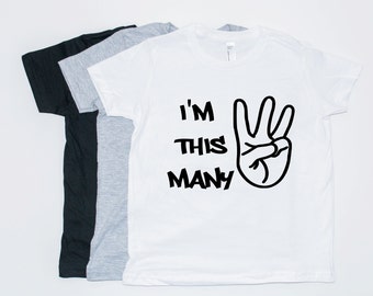 May include: White t-shirt with black text that reads "I'm this many" and a black peace sign with three fingers raised.