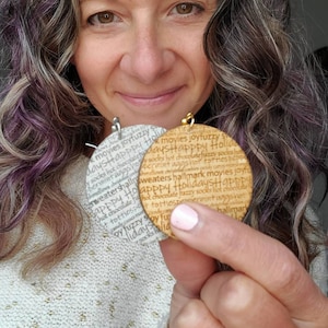 May include: A pair of round wooden earrings with a white background and gold lettering. The earrings feature a repeating pattern of words related to the holiday season, including "Happy Holidays", "fuzzy socks", "hot chocolate", and "movies".