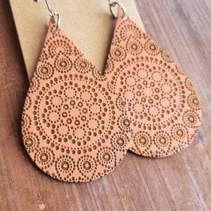 May include: A pair of peach-colored teardrop-shaped earrings with a mandala pattern. The earrings are made of wood and have silver hooks.