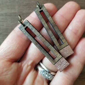 May include: A pair of wooden earrings with a geometric design. The earrings are brown with a rose gold accent. The earrings are hanging from a person's hand.