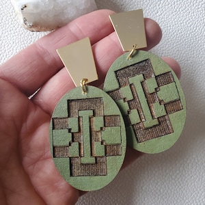 May include: A pair of green and gold oval-shaped earrings with a geometric design. The earrings are made of wood and have a gold metal backing.