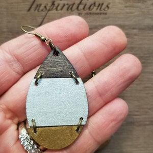 May include: A pair of teardrop-shaped wooden earrings with a brown, white, and gold color scheme. The earrings have a simple design with a single gold band separating each color.