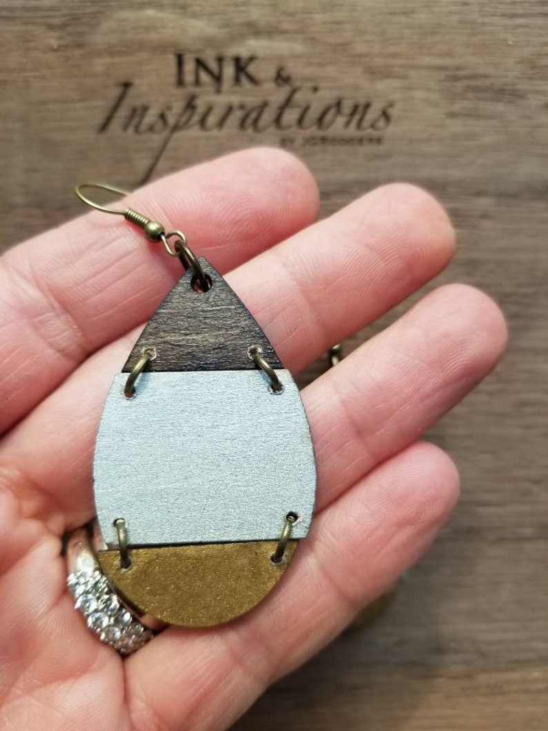 May include: A pair of teardrop-shaped wooden earrings with a brown, white, and gold color scheme. The earrings have a simple design with a single gold band separating each color.