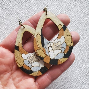 May include: A pair of wooden earrings with a floral design. The earrings are shaped like teardrops and have a black, white, and orange color scheme.