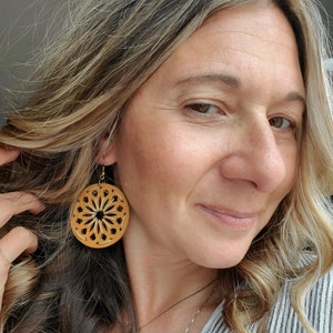 May include: A pair of round, brown leather earrings with a geometric design. The earrings are hanging from the earlobes of a person with long, wavy, light brown hair.