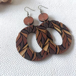 May include: A pair of dangle earrings featuring circular wooden components with a multi-tonal brown and tan leaf-like pattern. Each earring consists of a small solid circular top piece connected to a larger open-ring bottom piece with metal hooks.