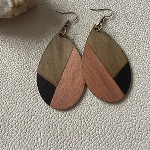 A pair of teardrop-shaped earrings lightweight wood earrings with a geometric block design. The earrings are made of thin wood and have a black, walnut stain and rose gold finish sections.