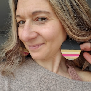May include: A pair of large, round earrings with a black background and colorful stripes. The stripes are pink, yellow, and white. The earrings are made of wood and have a natural finish.