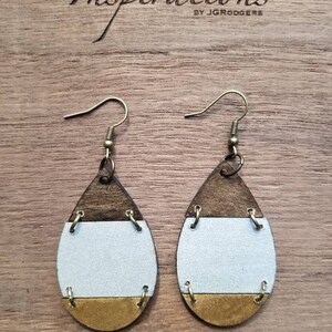 May include: A pair of teardrop-shaped earrings with a wood, white, and gold design. The earrings have a simple hook closure.