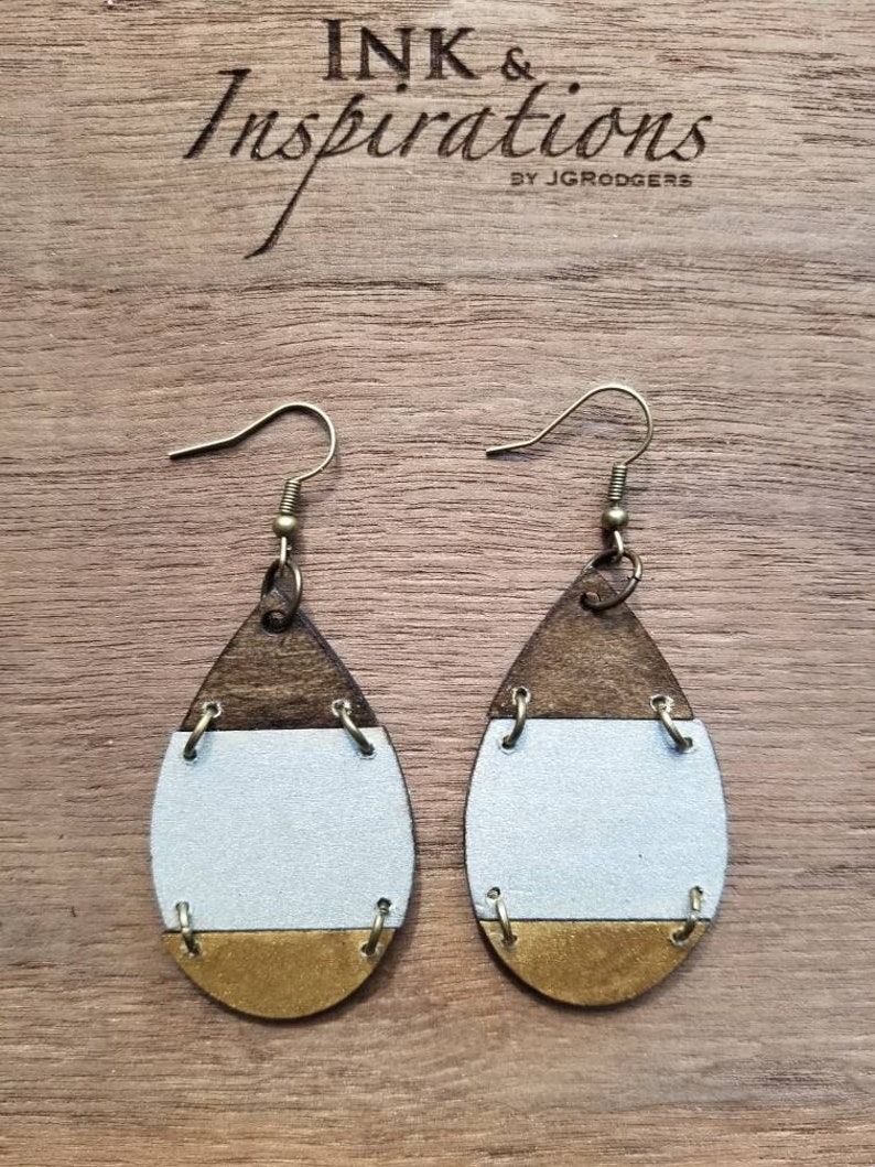 May include: A pair of teardrop-shaped earrings with a wood, white, and gold design. The earrings have a simple hook closure.