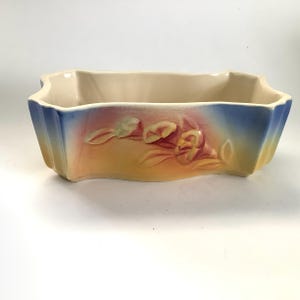 May include: A rectangular ceramic planter with a cream-colored interior and a gradient exterior. The exterior transitions from blue to orange and yellow, with embossed floral details. A decorative piece for plants.