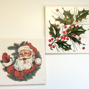 May include: Two square ceramic tiles with Christmas-themed designs. One tile features Santa Claus in a red suit and hat, surrounded by a green wreath. The other tile displays a sprig of holly with green leaves and red berries.