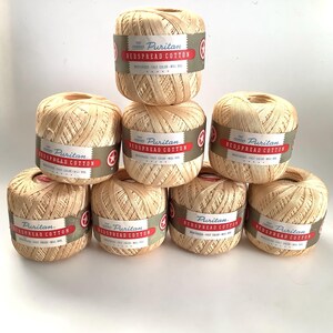 May include: Seven balls of light beige Puritan bedspread cotton yarn arranged in a pyramid stack. Each ball features a green and red paper label with the text The Famous Puritan Bedspread Cotton, mercerized, fast color, will boil.