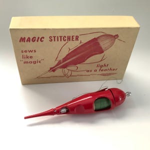 May include: A vintage red Magic Stitcher sewing tool with a cream-colored box. The box features the text "MAGIC STITCHER" and an illustration of the tool. The sewing tool is red and has a spool of green thread visible.