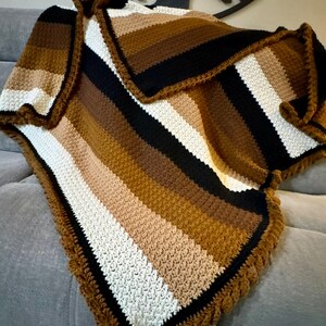 May include: A crocheted blanket featuring a repeating striped pattern in shades of black, white, tan, and dark brown. The blanket has a textured stitch throughout and is finished with a thick, ruffled border in a solid dark brown yarn.