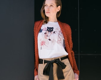 May include: A woman wearing a white t-shirt with a black and white graphic print of a panther, a rust-colored cardigan, and a beige skirt with a black bow.