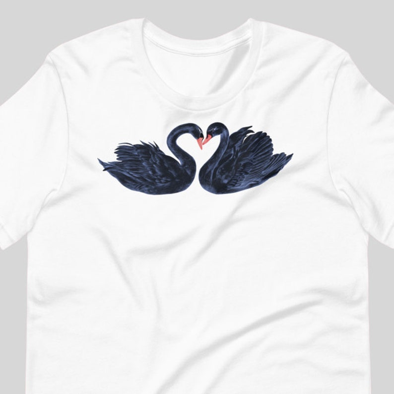 May include: A white t-shirt with a black and blue illustration of two black swans with their necks intertwined in a heart shape.