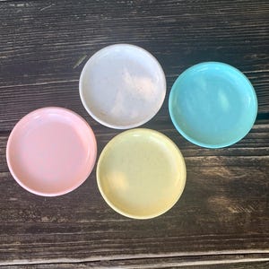 May include: Four small, round, ceramic dishes in pastel colors: pink, yellow, white, and blue. The dishes have a speckled texture and are arranged on a dark wooden surface. These dishes are suitable for serving small portions of food or as decorative items.