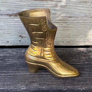 May include: A polished brass boot-shaped vessel featuring embossed details including a series of three circular eyelets near the top opening and horizontal lines along the shaft, with a curved heel and a pointed toe design.