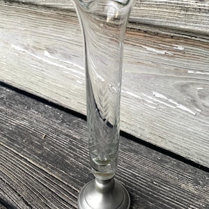 May include: A clear glass bud vase with a flared, ruffled top and a decorative etched pattern. The vase is mounted on a silver-colored metal base. The vase is approximately 8 inches tall.
