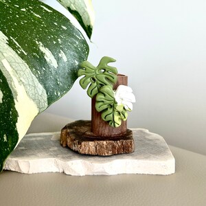 May include: A miniature wooden vase featuring a monstera leaf design. The cylindrical vase is set on a small wooden base. The leaves are green and white, creating a natural, botanical theme. The vase is approximately 5 cm tall.
