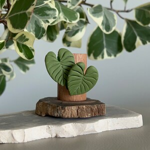 May include: Two green leaf-shaped polymer clay earrings on a wooden stand. The stand is on a white surface.