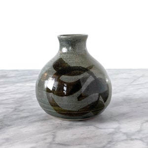 Può includere: Un piccolo vaso rotondo in ceramica grigia con un design astratto marrone. Il vaso ha un'apertura stretta e una base larga.