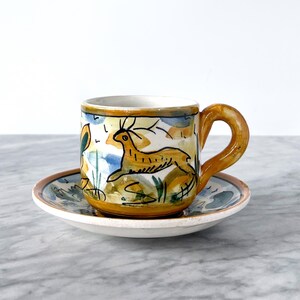 May include: A yellow and blue ceramic coffee cup with a painted design of a deer running through a field. The cup is sitting on a matching saucer.