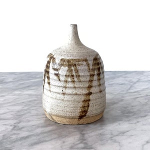 Könnte beinhalten: Eine kleine, weiße Keramikvase mit brauner Glasur und einer strukturierten Oberfläche. Die Vase hat einen runden Körper und einen schmalen Hals.