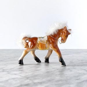Op de afbeelding: Een bruin en wit keramiek paardje met een witte manen en staart. Het paard draagt een geel en wit zadel met een parelrand.