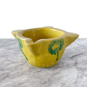 May include: A yellow ceramic mortar and pestle with green floral designs. The mortar is a small, shallow bowl with a wide rim and a rounded base. The pestle is a small, rounded stick with a handle.
