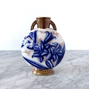 May include: A round, white ceramic vase with a blue floral design and gold accents. The vase features two small handles and a gold rim. The floral design includes blue lilies. The vase is approximately 6 inches tall.