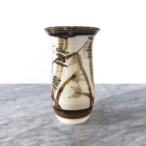 May include: A white ceramic vase with a brown and green floral design. The vase has a flared top and a rounded bottom.