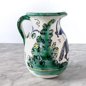 Könnte beinhalten: Handbemalter Keramikkrug mit weißem Sockel, grünem Rand und Griff. Der Krug hat ein skurriles Design mit grünem Laub und blauen Akzenten. Der Krug ist etwa 15 cm hoch.