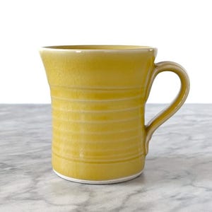 May include: A yellow ceramic mug with a curved handle. The mug has horizontal textured lines and a white base. The mug is set against a white background.