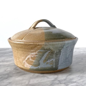 May include: A handmade ceramic lidded bowl with a handle. The bowl is a mix of tan, grey, and blue-green colours. The lid has a matching colour scheme. The bowl is sitting on a marble surface.