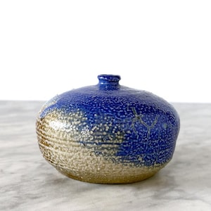 May include: A small, round ceramic vase with a textured surface. The top half is a deep blue, transitioning to a tan base. The vase has a narrow neck and a speckled pattern, creating a unique, handcrafted appearance. Ideal for displaying small flowers or as a decorative accent.