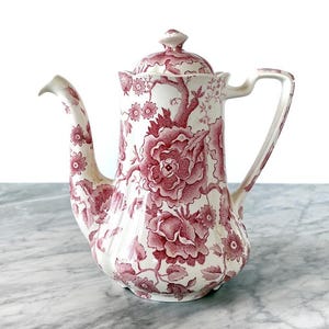 May include: A white ceramic coffee pot with a lid, handle, and spout. The pot is decorated with a red floral pattern. The pot is sitting on a marble surface.