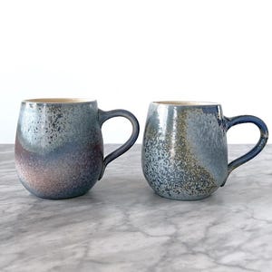 May include: Two ceramic mugs with a speckled, iridescent glaze in shades of blue, gray, and purple. The mugs have a rounded shape with a dark blue handle and a light-colored interior. They are placed on a marble surface.