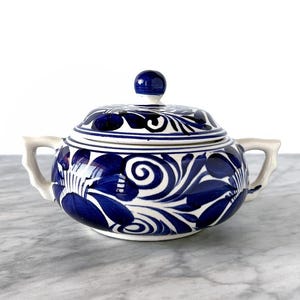 Op de afbeelding: Een blauw-witte keramische suikerpot met deksel, versierd met een bloemmotief. De pot heeft twee handvatten en een kleine knop op het deksel. Het ontwerp toont gestileerde bloemen en wervelende patronen.