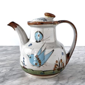 Könnte beinhalten: Eine Keramik-Teekanne mit cremefarbener Glasur, verziert mit handgemalten blauen Vögeln, einem Schmetterling und Blumenmotiven. Die Kanne hat einen braunen Rand, einen geschwungenen Henkel, einen braun akzentuierten Ausguss und einen passenden Deckel.